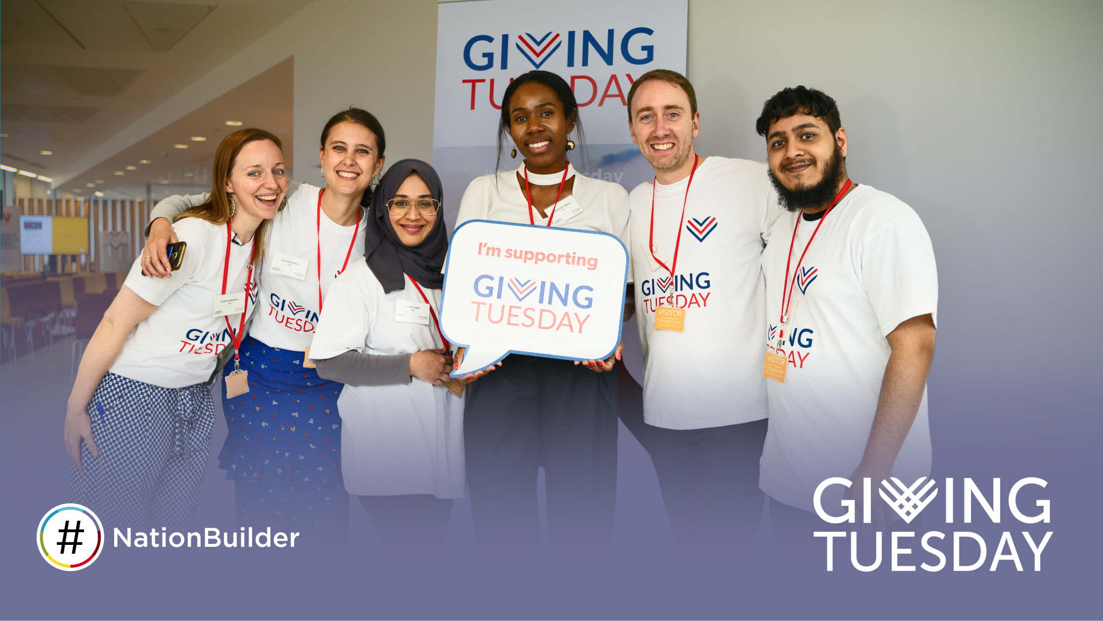 Six diverse volunteers stand together smiling in a bright office space, wearing white Giving Tuesday t-shirts and red lanyards. The person in the center holds a rounded square sign. A banner and logo appear in the background and corner.